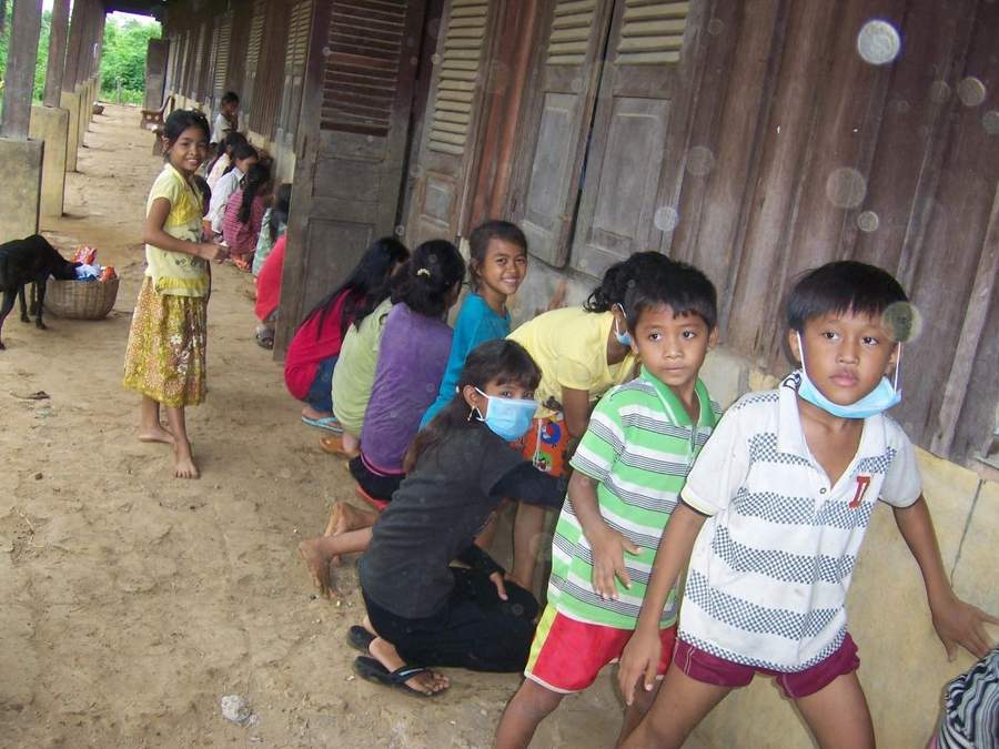 Children sitting along wooden building wall some with face masks