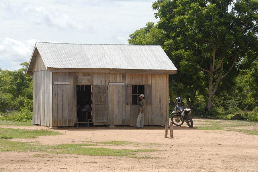 Wooden schoolhouse with tin roof and man standing outside with motorcycle